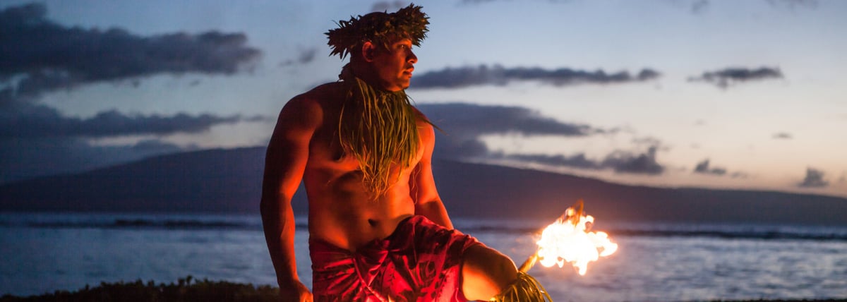 A luau performer in traditional costume at dusk, with warm stage light and ocean behind him.