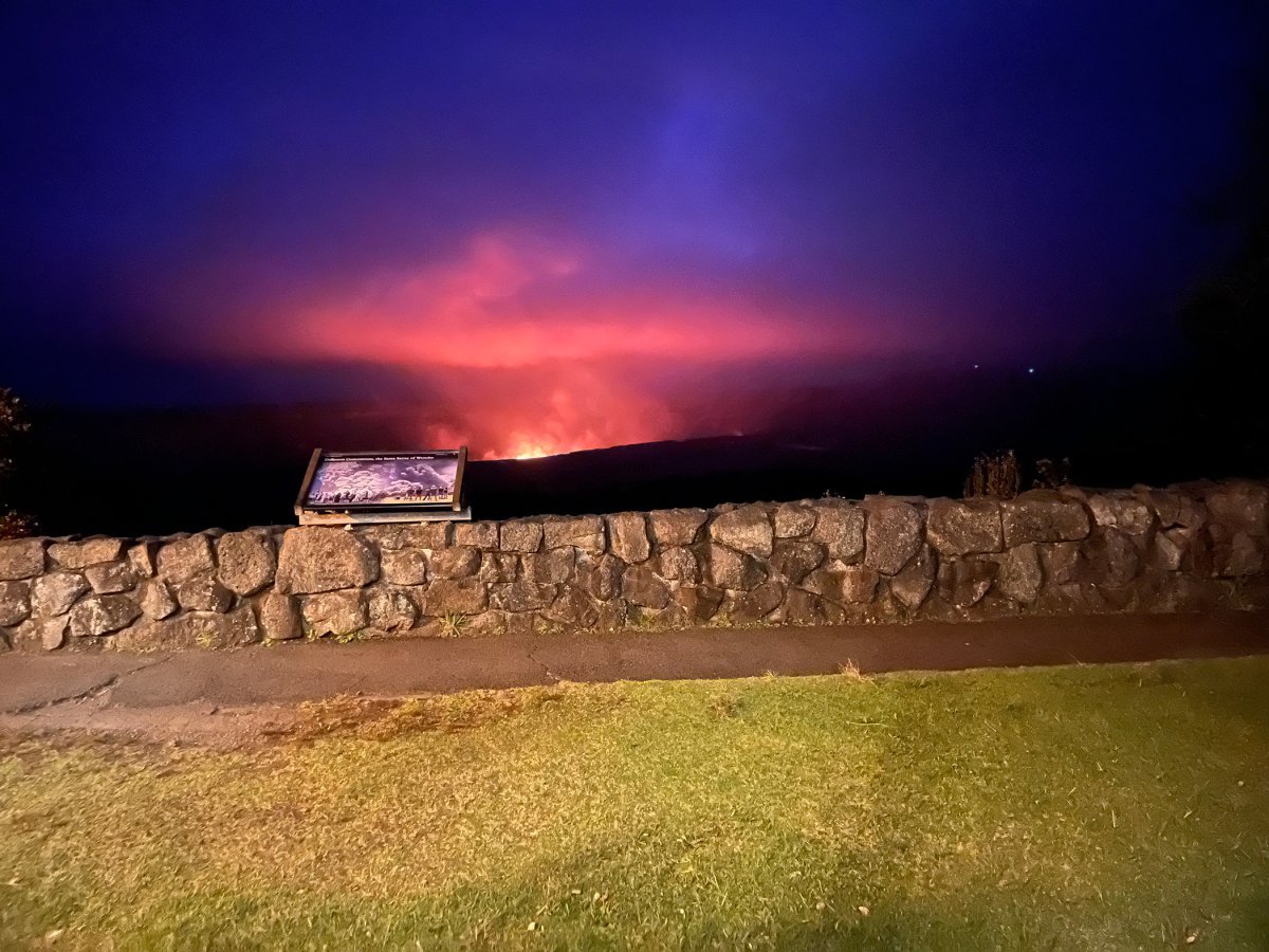 Nighttime view of Kīlauea Caldera glowing orange in the distance, seen from a lit restaurant patio inside Hawaiʻi Volcanoes National Park.