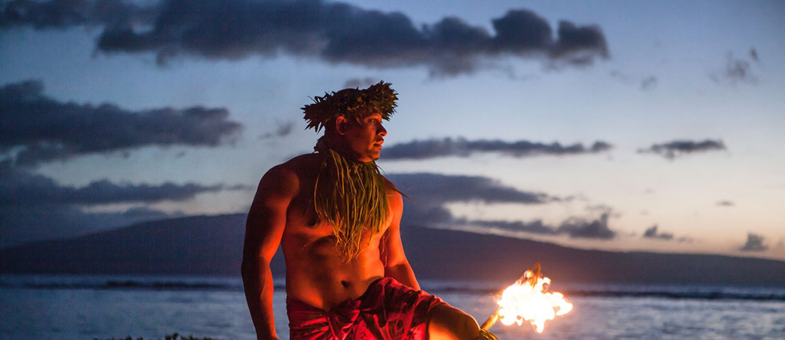 A fire dancer holding a burning torch at dusk along a shoreline, with the ocean and clouds behind him