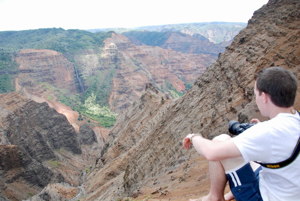 Waimea Canyon on Kauaʻi seen from the Puʻu Hinahina Lookout, with red-earth walls dropping thousands of feet and Waipoʻo Falls threading down the canyon face.