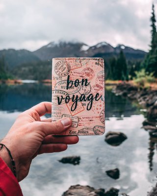 Person's hand holding up a Bon Voyage travel book with water and mountains in the background.