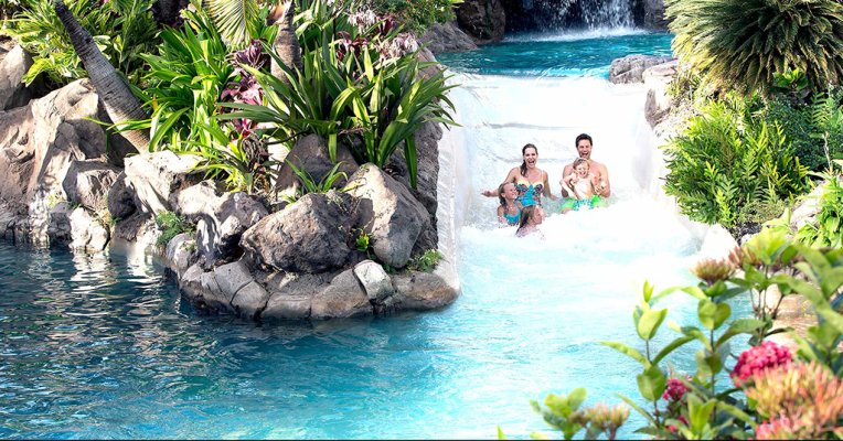 A family going down a water slide connecting two pools in a tropical setting at Grand Wailea Resort in Maui.
