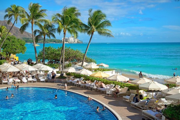 Several people swimming in a large circular pool with cabanas that overlooks the beach and ocean outside of the Halekulani in Oahu.
