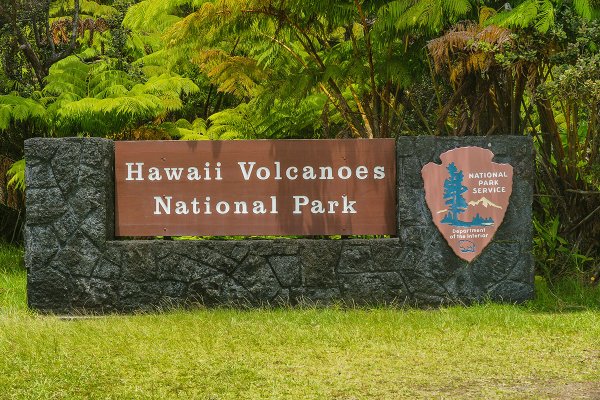 Hawaii Volcanoes National Park stone and brown sign outside of the entrance with vegetation in the background.
